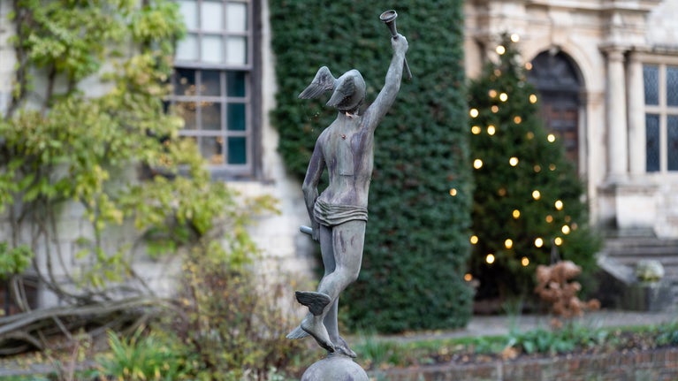 Wintry garden view with a statue in the foreground and twinkling lights on a Christmas tree close to a stone house beyond